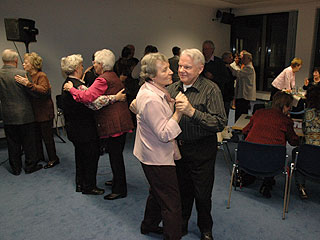 Im Vordergrund tanzen drei Paare, im Hintergrund ist geselliges Zusammensein an Tischen